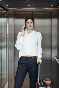 Businesswoman with suitcase taking on smart phone while standing in elevator