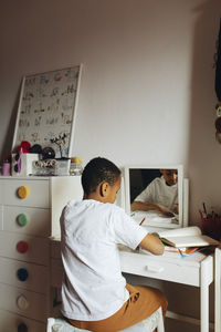 Rear view of boy writing in book while sitting at table in study room at home