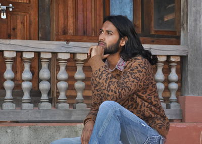 A attractive long haired indian young man looking sideways, posing with sitting on temple stairs