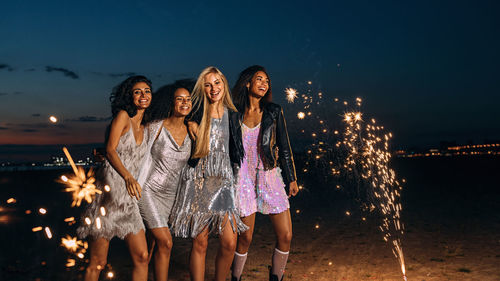 Cheerful young female friends with firework at beach against sky during dusk