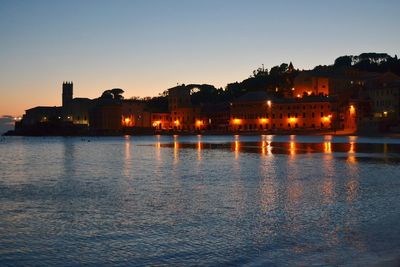 Illuminated buildings by river against sky at sunset