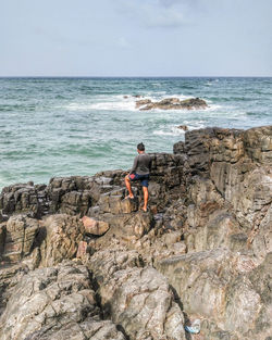 Rear view of man standing on rock at sea shore against sky