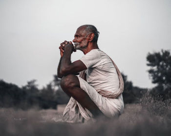 Side view of man sitting on field against sky