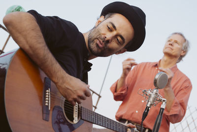 Portrait of young man wearing hat singing by microphone standing against sky