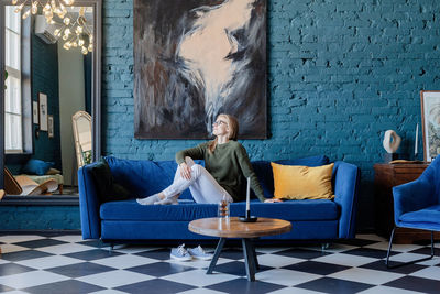 Young caucasian woman in green sweater and white jeans lying on sofa in stylish blue living room