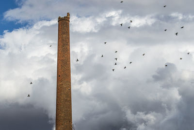 Low angle view of birds flying by damaged tower against cloudy sky