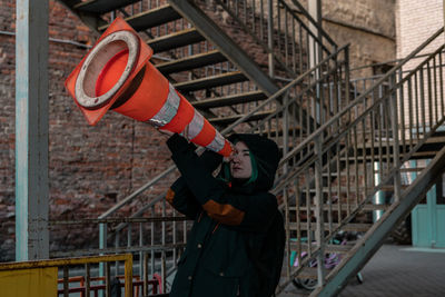 Side view of young woman looking through traffic cone while standing against building