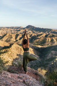 Back view of unrecognizable dark haired female standing on top of rocky mountain and doing tree with arms up pose