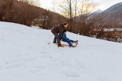 Father pushing daughter sitting on wooden sled to slide down the snowy hill
