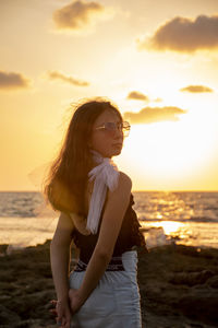 Young woman looking away while standing at beach against sky during sunset