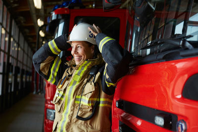 Smiling female firefighter wearing helmet while standing by fire engine at fire station