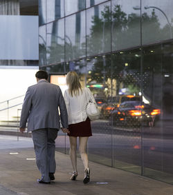 Full length rear view of couple walking by glass building