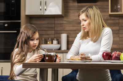White mom and little daughter talks while having breakfast in kitchen, girl holds cell phone, device