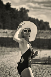Portrait of a woman in a swimsuit, hat and sunglasses in summer on the riverbank against the sky