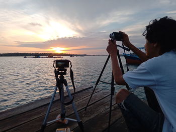 Man photographing at sea against sky during sunset