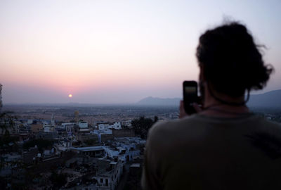 Rear view of woman photographing cityscape against sky during sunset