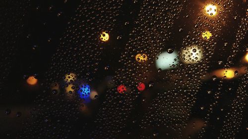 Illuminated lights on street seen through wet glass window at night