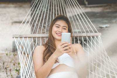 Smiling woman using phone while relaxing on hammock