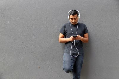 Young man listening music while using mobile phone against wall