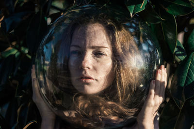 Close-up of young woman wearing glass ball on head against leaves