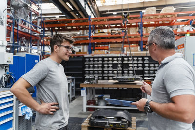 Smiling male coworker looking at mature engineer in illuminated factory