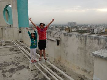 Portrait of happy siblings with arms raised standing on building terrace against cloudy sky