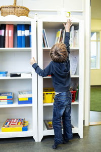 Rear view of boy reaching for container on shelf in kindergarten