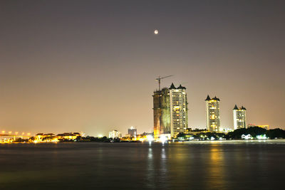 Illuminated buildings by river against sky at night