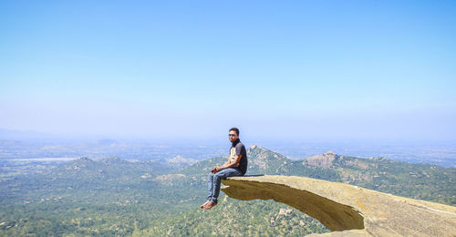 Man sitting at the edge of cliff against clear blue sky