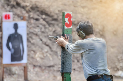 Rear view of man holding gun while aiming on target outdoors