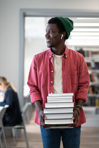 Cheerful positive african american man with stack of books walks through library enjoying studying
