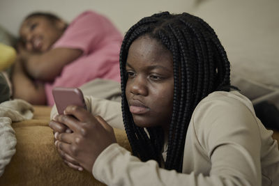 Teenage girl using smart phone by female friend lying in bedroom at home