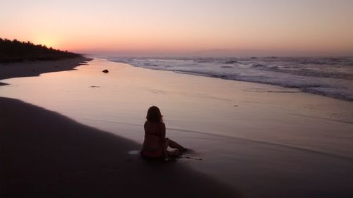 Man sitting on shore at beach against sky during sunset