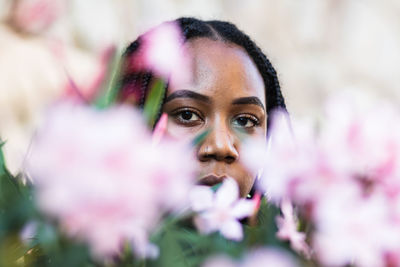 Soft focus of young african american woman with braids looking at camera from behind natural pink flowers