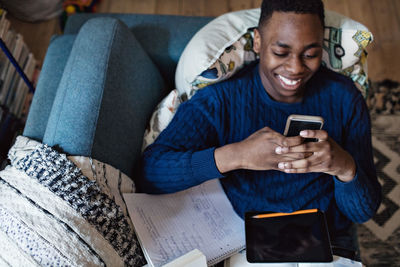 High angle view of social media addicted teenage boy using smart phone while lying on sofa with books in living room