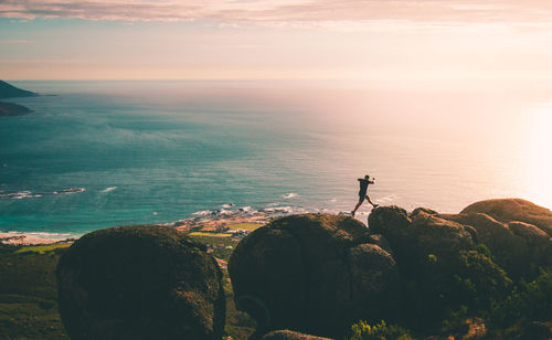 Man jumping on rock at beach against sky during sunset