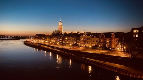 Illuminated buildings by river against sky at dusk