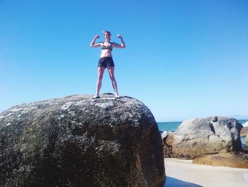 Low angle view of woman flexing muscles while standing on rock against sky 