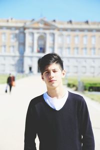 Portrait of young man standing on footpath against building during sunny day