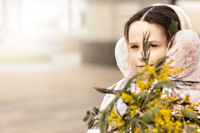 Portrait of a toddler girl in a warm pink coat and fur headphones with a bouquet of mimosa 