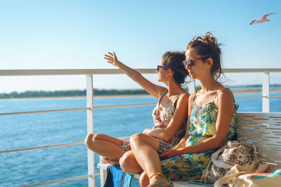 Women sitting on seat by railing against sea