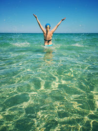 Rear view of woman with arms raised standing in sea against clear blue sky during sunny day