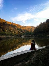 Rear view of young woman sitting by lake against sky during autumn