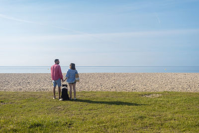 Back view of couple standing and holding hands near their dog on beach