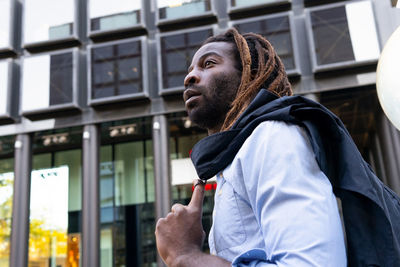 Young bearded black male with dreadlocks wearing white shirt holding the jacket over the shoulder while looking away in modern office building