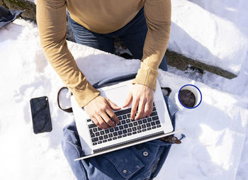 Mid adult man using laptop by coffee cup kept on snow covered bench in sunlight