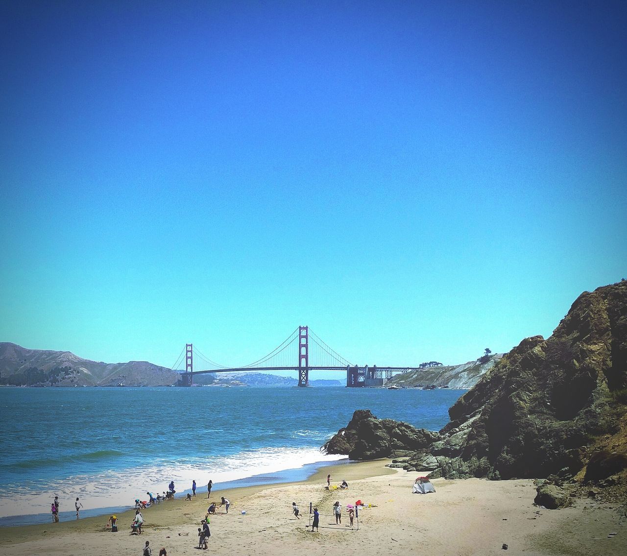 clear sky, copy space, water, large group of people, built structure, sea, beach, blue, architecture, connection, suspension bridge, tourism, tourist, leisure activity, bridge - man made structure, travel destinations, vacations, famous place, engineering