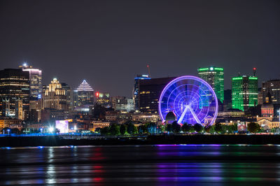 Illuminated ferris wheel by river against buildings at night