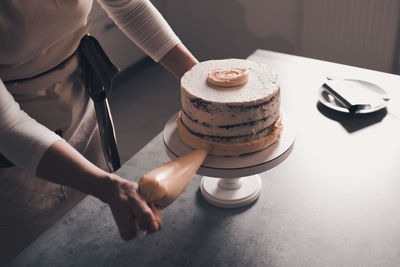 Woman decorating cake with whipped cheese cream holding pastry bag closeup in kitchen.