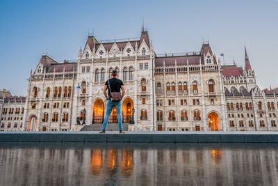 Rear view of man standing on land by water against building and clear sky